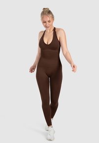 Brun, tætsiddende jumpsuit med dyb V-halsudskæring og tynde stropper, lavet af et glat, elastisk materiale. Bruges med hvide sportssko.