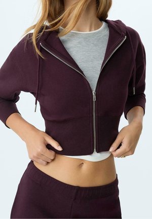 Frau trägt einen kurzen, dunkelvioletten Hoodie mit Reißverschluss über einem grauen Top, zeigt ihren Bauch und zieht am Reißverschluss des Hoodies.