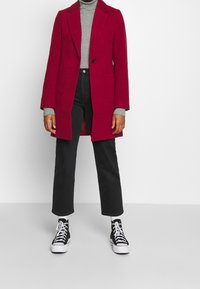 Abrigo largo rojo con cuello con muesca, cierre de un solo botón y bolsillos laterales, combinado con un suéter de cuello alto gris, jeans negros y zapatillas.