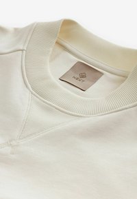 Sudadera beige claro con cuello redondo acanalado, costuras planas y una etiqueta rectangular de cuero con la marca "NEXT".