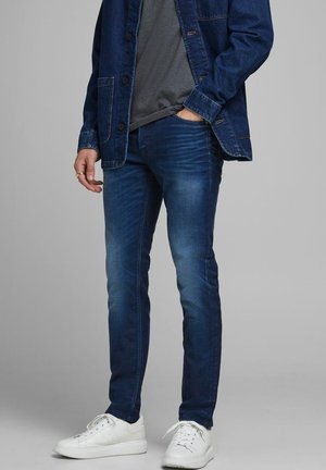 Vaqueros de mezclilla azul oscuro con corte slim, detalles descoloridos, combinados con una camiseta gris y una chaqueta azul, complementados con zapatillas blancas.