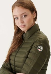 Olijfgroen gewatteerde jas met een hoge kraag en ritssluiting. Voorzien van een ronde logopatch op de linkermouw. Geweven, geborduurd ontwerp.