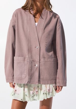 Femme portant une veste mauve boutonnée avec de grandes poches devant par-dessus une jupe légère à volants fleurie.