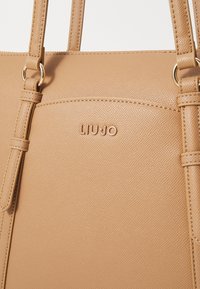 Borsa a tracolla tan realizzata in materiale sintetico testurizzato, con due manici, caratterizzata da un logo impresso e accenti in metallo dorato.