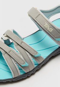 Teva sandals featuring gray textile straps, a light blue cushioned insole, and a textured black rubber outsole. Adjustable strap design.