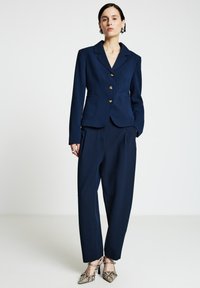 Blazer bleu marine avec boutons dorés, deux poches avant, assorti à un pantalon coordonné. Des talons texturés à motif serpent ajoutent une touche à la tenue.
