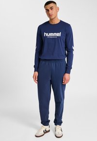 Granatowa koszula z długim rękawem z białym logo "hummel", zestawiona z pasującymi granatowymi spodniami dresowymi i kremowymi sneakersami z czarnymi akcentami.