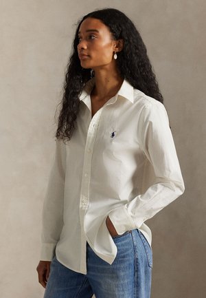 Kvinde med langt krøllet hår iført hvid skjorte med knapper og logo samt blå jeans står med den ene hånd i lommen og kigger mod venstre.