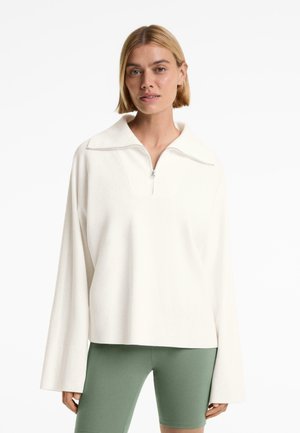 Pull blanc en maille avec col zippé, manches larges et poignets côtelés, associé à un short de vélo vert olive. Texture lisse.