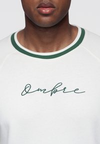 Vit sweatshirt med en grön ribbad krage och broderad text "Ombre" i kursiv stil. Mjuk textur, avslappnad design.