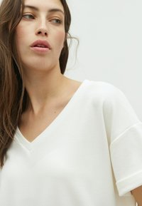 Camiseta blanca de cuello en V con mangas cortas y enrolladas, hecha de una tela suave, que presenta un corte relajado y una textura lisa. Diseño sencillo sin patrones.