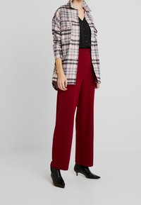 Camicia a quadri in misto lana nei toni del rosa, grigio e nero, indossata sopra una maglietta nera, abbinata a pantaloni larghi a vita alta bordeaux e stivali neri alla caviglia.