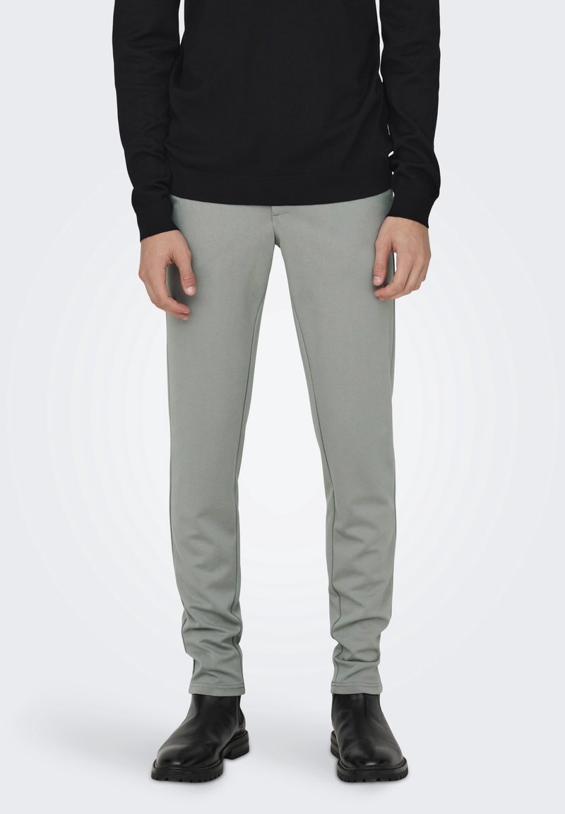 Pantaloni slim-fit grigi chiari realizzati in tessuto morbido, con una texture liscia e vita elastica, abbinati a stivaletti neri alla caviglia.