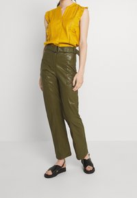 Žlutá volánková blůza s detaily knoflíků, spárovaná s olivově zelenými kalhotami s vysokým pasem z umělé kůže a černými plochými sandály se stuhami.