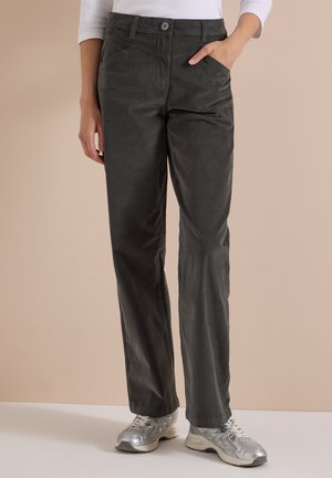 Personne portant un pantalon droit gris foncé avec poches avant, un haut blanc à manches longues, et des baskets argentées sur un fond uni.