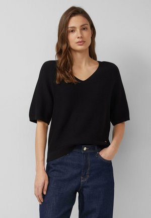 Junge Frau mit braunen Haaren trägt einen schwarzen Kurzarm-V-Ausschnittpullover und dunkelblaue hoch taillierte Jeans, steht vor einem schlichten hellgrauen Hintergrund.