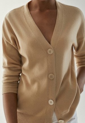 Femme portant un cardigan beige boutonné avec poignets et ourlet côtelés, manches retroussées, une main dans la poche.