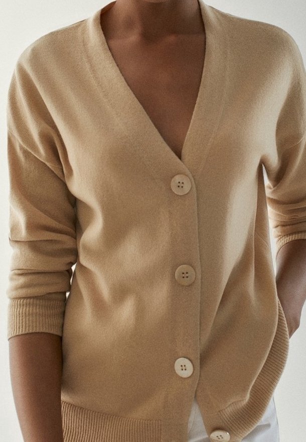 Femme portant un cardigan beige boutonné avec poignets et ourlet côtelés, manches retroussées, une main dans la poche.