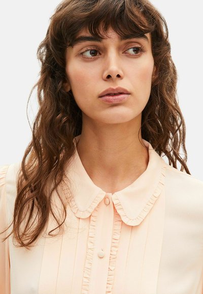 Joven con cabello castaño ondulado que lleva una blusa color durazno claro con un cuello Peter Pan con volantes, mirando ligeramente hacia un lado.