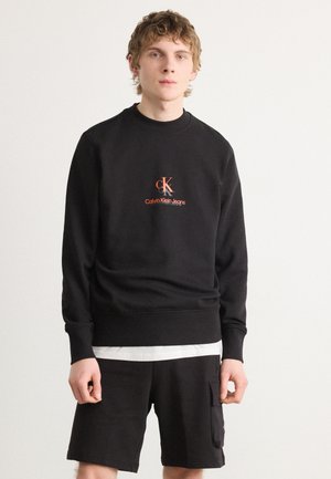Sweatshirt noir avec des poignets et un ourlet côtelés, comportant un logo brodé orange "ck Calvin Klein Jeans." Porté avec un short noir assorti.
