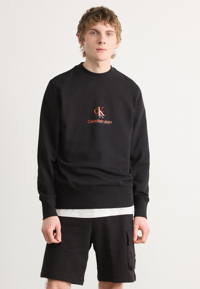 Felpa nera con polsini e orlo a costine che presenta un logo ricamato arancione "ck Calvin Klein Jeans." Indossata con pantaloni corti neri coordinati.