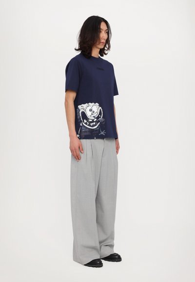 Marineblå bomulds T-shirt med tegneserieprint og rund hals, parret med lysegrå plisserede bukser og sorte sko, mod en ensfarvet baggrund.