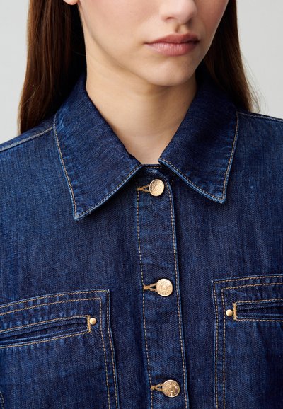 Chaqueta de denim de tono azul oscuro, con un cuello puntiagudo, botones en tono dorado y dos bolsillos en el pecho con detalles de costura.