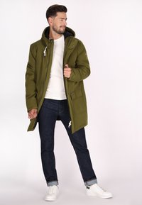 Giacca parka verde con chiusura a zip, tasche con patta e cappuccio con cordino. Indossata sopra una t-shirt bianca, abbinata a jeans scuri e sneaker bianche.