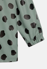 Vandafvisende grøn jakke med et sort uregelmæssigt prikmønster, elastiske manchetter og en glat, blank overflade.