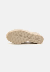 Suela de sandalia de plataforma beige con agarre texturizado y líneas horizontales elevadas. Logo "Calvin Klein Jeans" estampado cerca del talón.