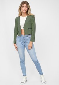 Groene korte jas met een gladde textuur, voorzien van lange mouwen en een voorkant ritszak, gecombineerd met lichtblauwe skinny jeans en witte sneakers.