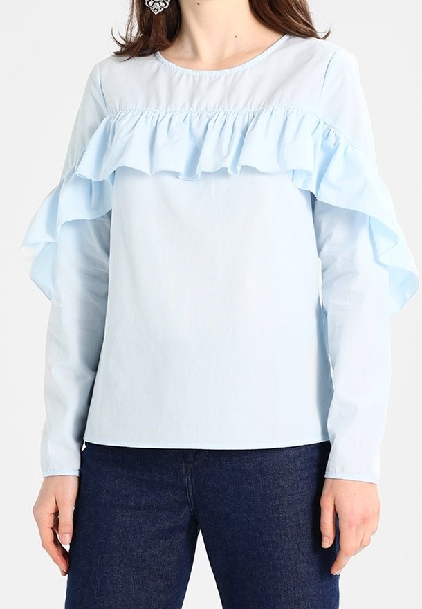 Blouse bleu clair à manches longues avec un détail à volants sur la poitrine et les manches, portée avec un jean bleu foncé.