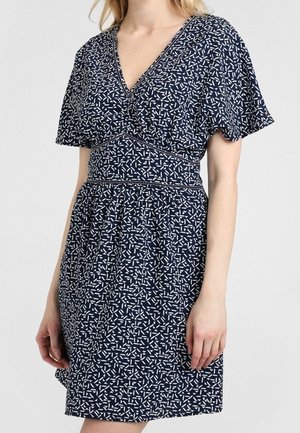 Frau trägt ein marineblaues Kleid mit weißen abstrakten geometrischen Mustern, kurzen flatternden Ärmeln und einem Taillendetail mit Raffungen.