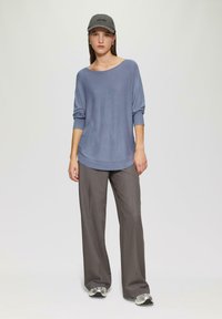 Hellblaues, langärmliges Oberteil mit weitem Ausschnitt, kombiniert mit grauen, weiten Hosen und silbernen Sportschuhen. Eine graue Mütze rundet das Outfit ab.