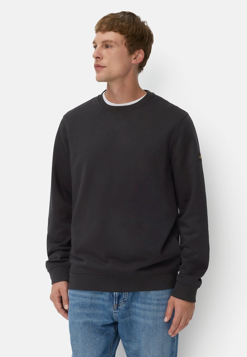 Donkergrijze crewneck sweatshirt met lange mouwen, ribgebreide boorden en zoom. Heeft een klein logo op de linkerarm. Draag het over een wit T-shirt.