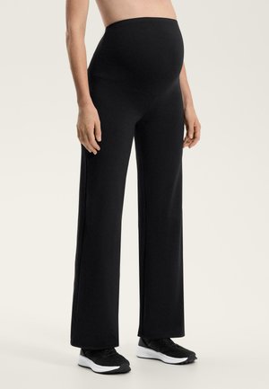 Terhes személy fekete bő szárú kismama nadrágot és fekete tornacipőt visel, egyszínű világos háttér előtt áll.