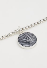 Bracelet en chaîne argentée avec un pendentif rond présentant une surface texturée gris foncé, mettant en valeur des motifs complexes et une bordure polie.