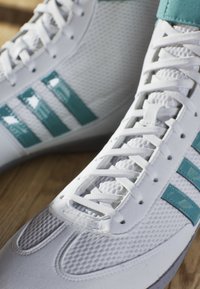 Bílé sportovní boty s tyrkysovými akcenty, texturovaný síťovaný materiál, ploché tkaničky a tři lesklé pruhy po stranách.