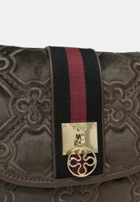 Bruine quilted handtas met een verhoogd geometrisch patroon, gouden hardware, een sluiting met een decoratief embleem en een zwart-rode gestreepte band.