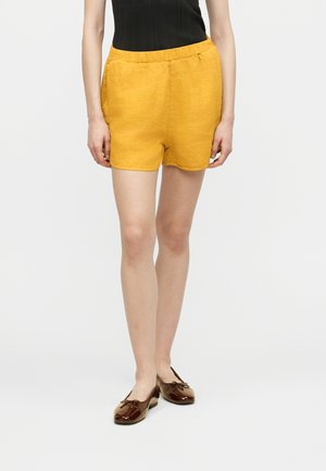 Nainen, joka pukeutunut sinappikeltaisiin pellavashortseihin, mustaan hihattomaan ribbineuletoppiin ja ruskeisiin kiiltonahkaisiiin balettitennareihin, seisoo yksiväristä taustaa vasten.