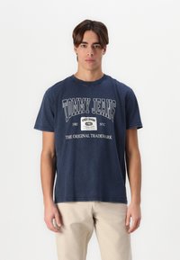 Tamsiai mėlynas medvilninis marškinėliai su baltais "TOMMY JEANS" tekstu ir logotipu. Laidena forma, trumpomis rankovėmis ir apvalia iškirpte.