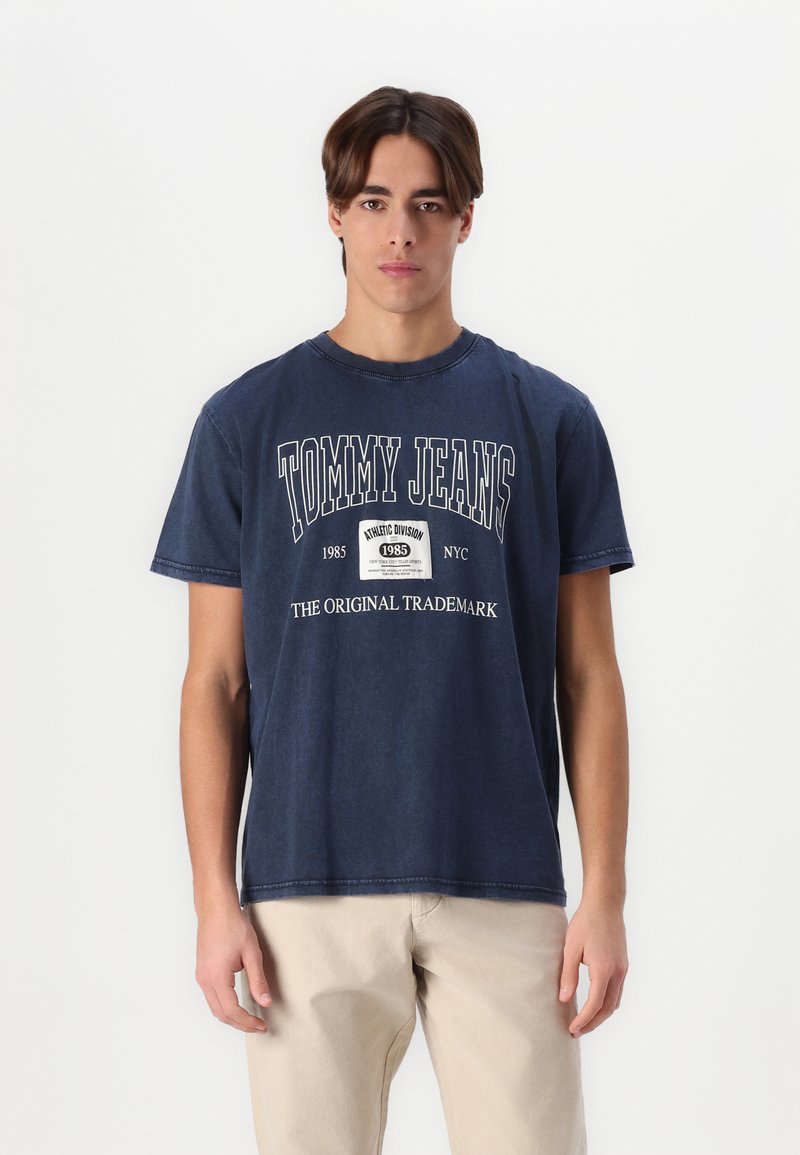 Tamsiai mėlynas medvilninis marškinėliai su baltais "TOMMY JEANS" tekstu ir logotipu. Laidena forma, trumpomis rankovėmis ir apvalia iškirpte.
