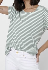 Camiseta de manga corta a rayas verdes y blancas con escote redondeado, hecha de material suave. Combinada con jeans blancos con acentos de botones dorados.