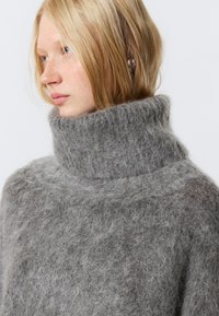 Pulover gri, supradimensionat, cu guler înalt, realizat dintr-un material moale și pufoasă. Are un guler gros și un design minimalist, fără modele.