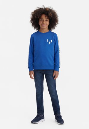 Barn med krøllet hår iført en blå langærmet skjorte, mørke jeans og mørke sneakers, står med et neutralt udtryk på en hvid baggrund.