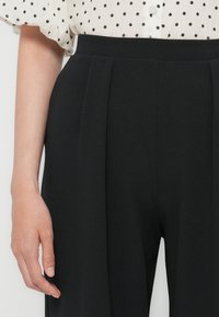 Pantalons noirs ajustés avec des plis, présentant une taille haute et une texture lisse. Une main reposant sur le côté ajoute du contexte.