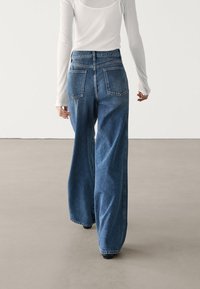Persona che indossa jeans azzurri a vita alta e gamba larga e una blusa bianca attillata a maniche lunghe, che cammina su un pavimento grigio liscio.