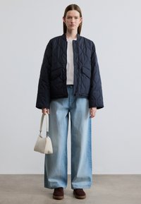 Καπιτονέ ναυτικό μπουφάν με μεγάλες τσέπες, σε συνδυασμό με ανοιχτό μπλε τζιν παντελόνι με φαρδιές μπατζάκια και καφέ αθλητικά παπούτσια. Κρατά μια κρεμ τσάντα χειρός.