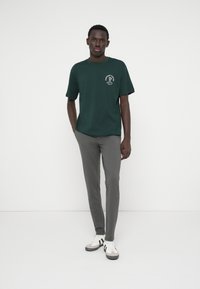 Tume roheline, lühikeste varrukatega puuvillane T-särk, millel on valge graafika vasakul rinnal, koos kitsaste hallide pükstega ja valgete spordijalatsitega.