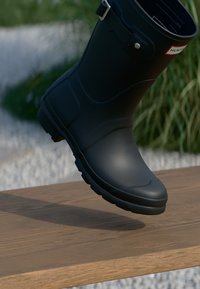 Botte en caoutchouc noire avec un orteil arrondi, une semelle texturée et une sangle ajustable. Posée sur une surface en bois avec de l'herbe verte en arrière-plan.